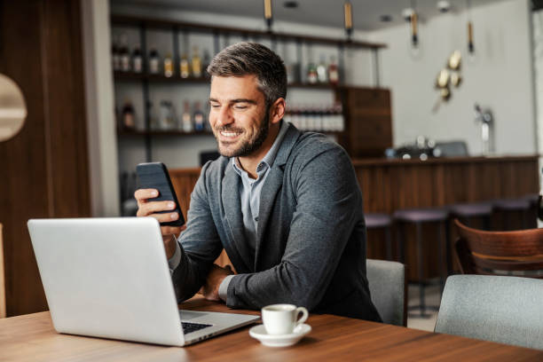 A happy businessman is sitting in a coffee shop and checking on his bank account on the mobile. There is a laptop on a table. A man using the phone for e-banking stock photo
