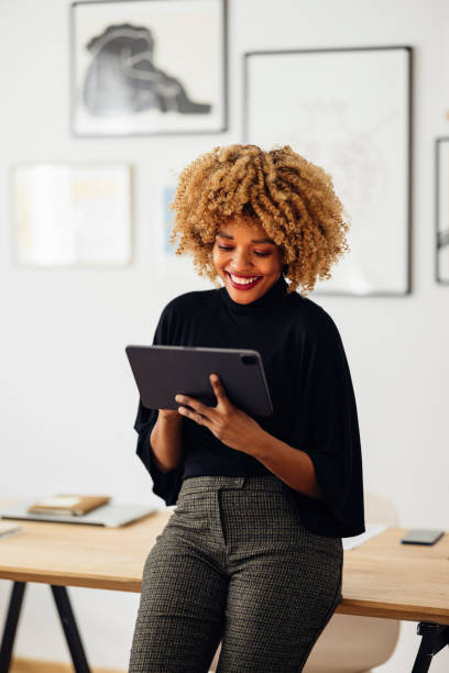 Happy Businesswoman Sitting on Her Desk Using Her Tablet Cheerful African American woman, sitting on her desk, smiling and using her laptop. She is typing something. coworking photos stock pictures, royalty-free photos & images