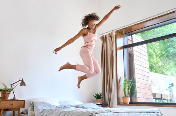 Happy carefree young African American woman wearing pajamas jumping on bed. stock photo