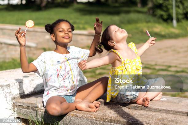 Happy caucasian and African-American girls with lollipops in their hands sitting in the lotus position on a summer day in the park and enjoying the sun.Summer concept,diverse people concept