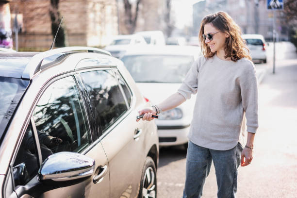 Happy cheerful young woman driver  in sunlasses opens her car with a remote key on the street in the city stock photo