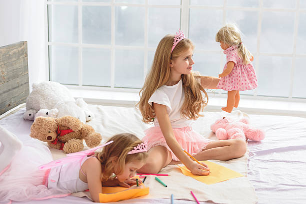 Happy children creating images in house Future artists. Good-looking girls drawing different pictures. Elder sister is painting her doll and smiling. Toys are on the big bed doll stock pictures, royalty-free photos & images