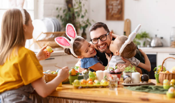 Happy children hugging and kissing father in kitchen Loving cute children wearing funny bunny ears headbands embracing and kissing cheerful young father while gathering with family at table in kitchen for Easter eggs painting easter stock pictures, royalty-free photos & images