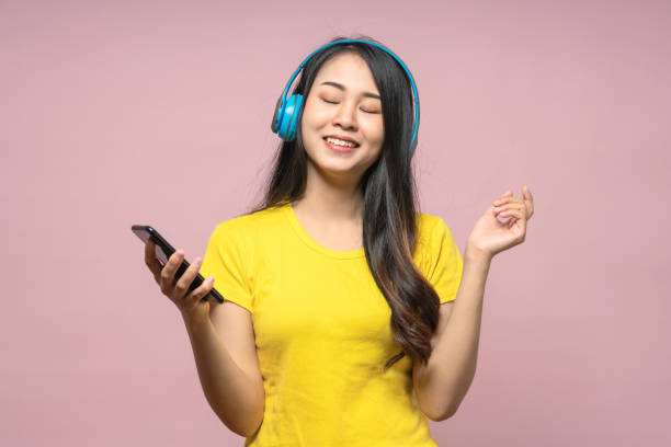 Happy chill asian girl listening to music in wireless headphone with smartphone. Woman listening podcast and wearing yellow shirt on pink isolated background. Happy chill asian girl listening to music in wireless headphone with smartphone. Woman listening podcast and wearing yellow shirt on pink isolated background. indonesian woman stock pictures, royalty-free photos & images