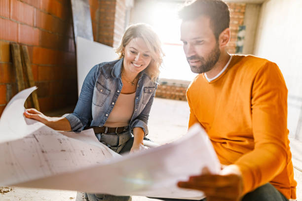 Happy couple analyzing plans at their renovating apartment. Young happy couple examining blueprints during home renovation process in the apartment. Focus is on woman. renovation stock pictures, royalty-free photos & images