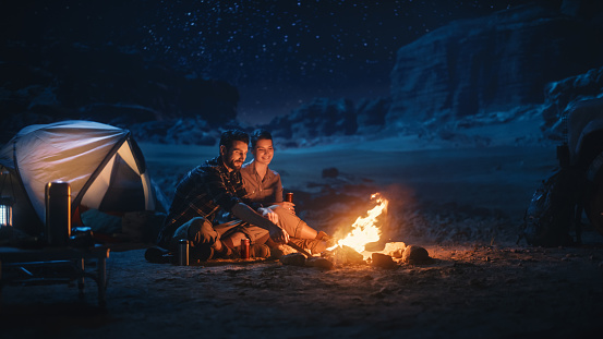 Happy Couple Nature Camping in the Canyon, Sitting Watching Campfire Together, Talking, Watching Night Sky. Two Traveling Young people On Inspirational Vacation Trip Marvel at Milky Way Stars