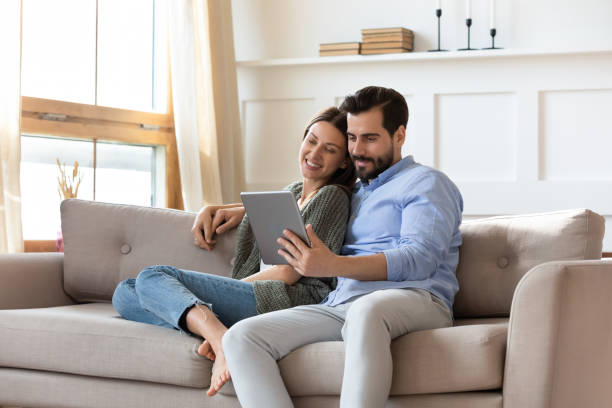 Happy couple relax on couch using tablet together Happy young couple sit relax on couch in living room watch movie on tablet together, loving millennial husband and wife rest on cozy sofa at home using modern pad gadget, family weekend concept two parents stock pictures, royalty-free photos & images