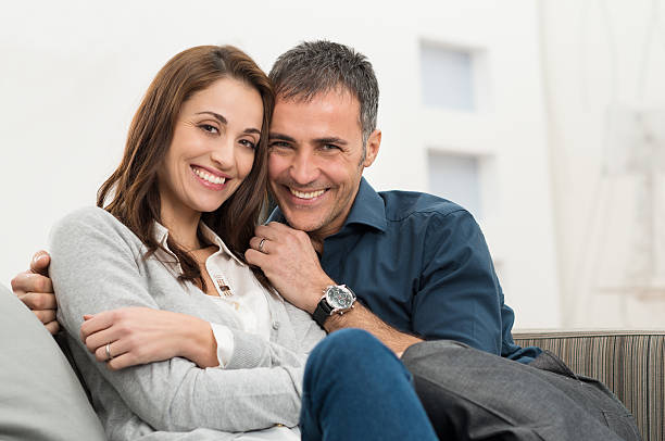 Happy Couple Sitting On Couch Happy Couple Embracing Sitting On Couch Looking At Camera mid adult couple stock pictures, royalty-free photos & images