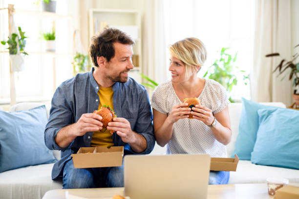 Happy couple sitting on sofa indoors at home, eating hamburgers. Front view of happy couple sitting on sofa indoors at home, eating hamburgers. take out food stock pictures, royalty-free photos & images