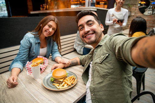 Happy Latin American couple taking a selfie while eating burgers at a restaurant   to post on their social media - lifestyle concepts