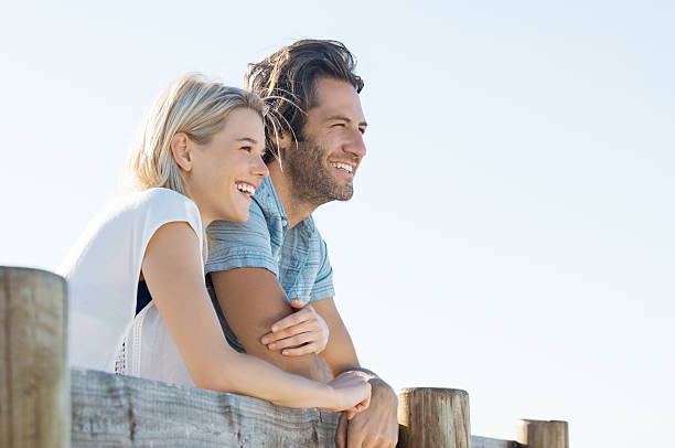 Happy couple vision Happy young couple leaning over fence and looking away, copy space. Smiling couple thinking about the future. Young woman embracing her boyfriend outdoor during the sunshine. mid adult couple stock pictures, royalty-free photos & images