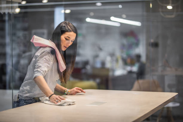 Happy creative woman cleaning the table before a meeting in the office. Smiling freelance worker cleaning the desk in the office. The view is through glass. clean desk stock pictures, royalty-free photos & images