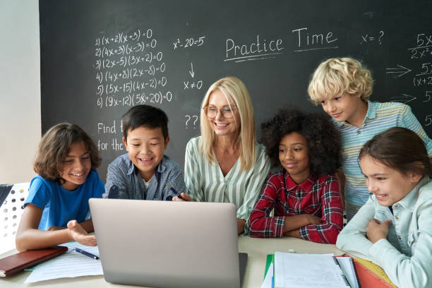 Happy diverse school children students gather at teacher table look at laptop. stock photo