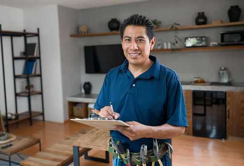 Happy Latin American electrician working at a house and looking at the camera smiling while writing on a clipboard - home improvement concepts