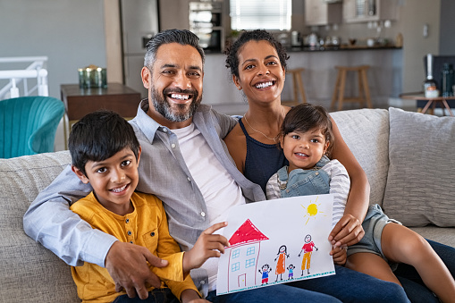 Proud parents showing family painting of son and daughter sitting on sofa at home. Smiling african mother and middle eastern father with two children looking at camera. Little boy and cute girl with parents showing handmade painting of a happy united family in a new home.