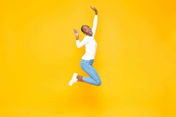 Happy excited African American woman jumping with hands raising in yellow isolated studio background with copy space stock photo