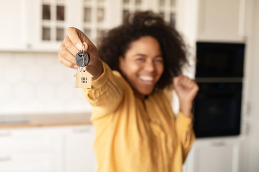 Joyful African-American woman holds keys with trinket in shape of little cute house, selective focus on the keys
