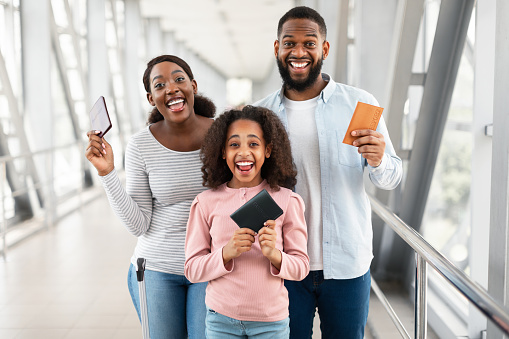 Portrait Of Cheerful African American Family Of Three People Holding Showing Passports And Tickets, Looking At Camera. Excited Mom, Dad And Daughter Posing Standing Indoors In Modern Airport Terminal