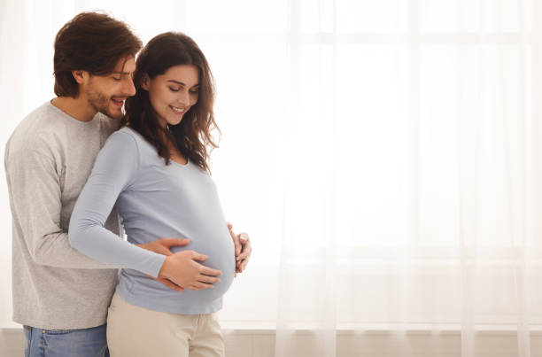 Happy expecting couple hugging while standing near the window at home Future parents. Happy expecting couple hugging and smiling while standing near the window at home, free space pregnant stock pictures, royalty-free photos & images