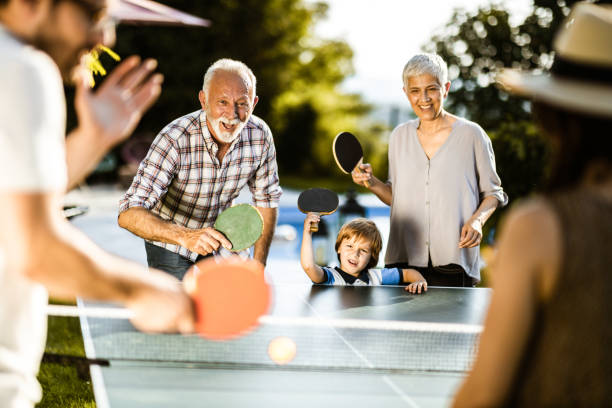 Happy extended family having fun while playing table tennis in the backyard. Happy grandparents and their small grandson having fun while playing table tennis against boy's parents in the backyard. table tennis stock pictures, royalty-free photos & images