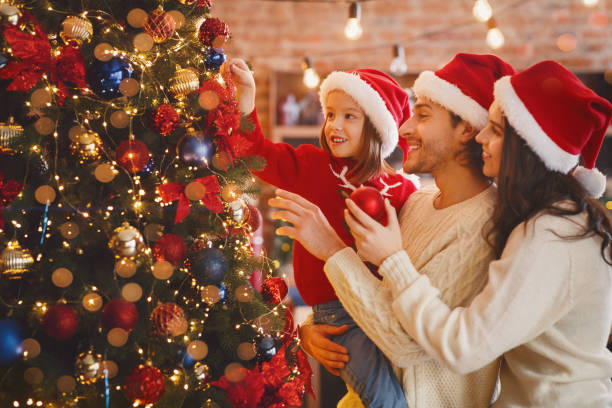 Happy family decorating xmas tree with bubbles at home Happy family of three in red Santa hats decorating xmas tree with bubbles and lights at home decorating photos stock pictures, royalty-free photos & images