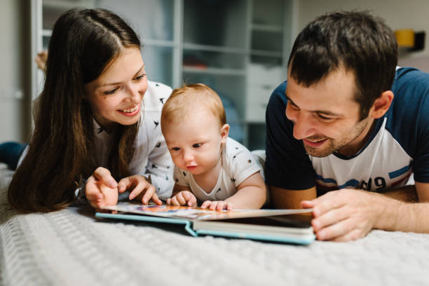 Happy family, parenthood and people concept - mother, father with baby lying in bed at home. Portrait of young smiling family with son reading book. stock photo