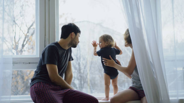 Happy family with young cute daughter sitting on windowsill playing and looking in window at home stock photo