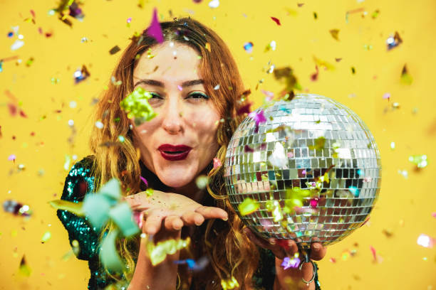 happy fashion girl blowing confetti from hands holding a disco ball with yellow background - young woman having fun at fest wearing trendy dress - party, event and celebration concept - focus on face - balo~es festa imagens e fotografias de stock