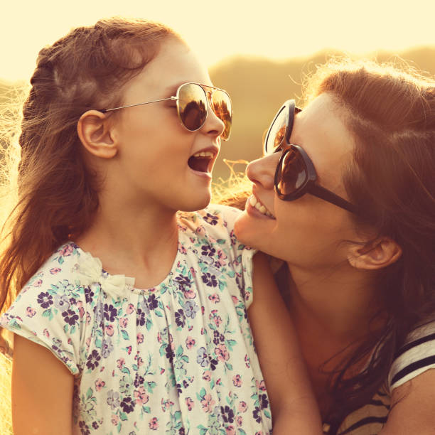 Happy fashion kid girl speaking to her mother in trendy sunglasses on nature sunset background. Closeup portrait of happiness. stock photo
