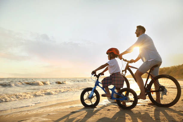 happy father with son riding bicycles on sandy beach near sea at sunset - fietsen strand stockfoto's en -beelden