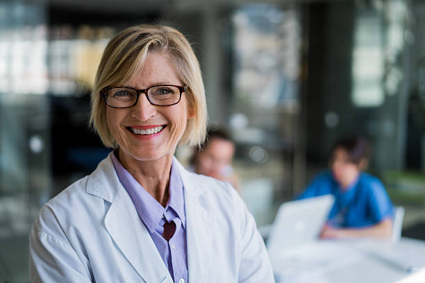 Happy female doctor in hospital A photo of confident female doctor in hospital. Portrait of happy mature professional wearing lab coat. Smiling expert is with colleagues working in background. lab coat stock pictures, royalty-free photos & images