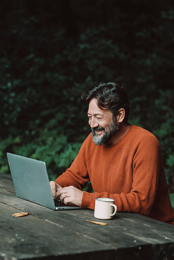 Happy free adult man work outdoors on a table with nature in background - concept of digital nomad modern online lifestyle people - mature male with beard use laptop computer in the woods smiling and enjoying