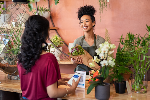 Young african florist holding beautiful bouquet of fresh flower while customer making online payment with smartphone while scanning QR code. Black saleswoman wearing apron giving a bunch of flower to customer at botany shop. Young woman making digital payment by phone wallet at flower shop.
