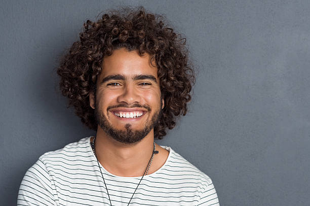 Happy friendly guy Portrait of a happy cheerful young man looking at camera. Handsome young man with beard and curly hair standing against grey background. Close up face of multi ethnic young man isolated against grey wall. curly hair stock pictures, royalty-free photos & images