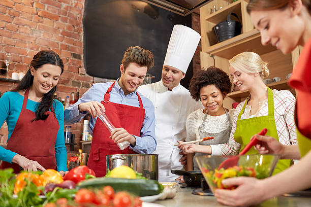 happy friends and chef cook cooking in kitchen cooking class, culinary, food and people concept - happy group of friends and male chef cook cooking in kitchen cooking class stock pictures, royalty-free photos & images