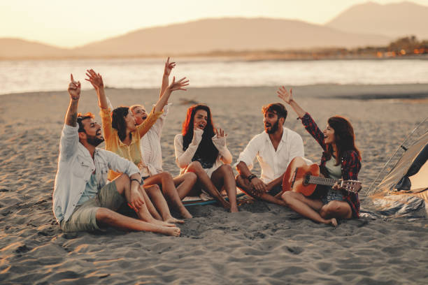 Happy friends sitting on the beach singing and playing guitar during the sunset stock photo