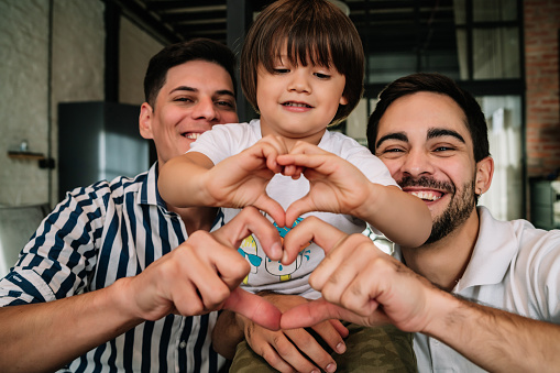 Happy gay couple posing with their son while making a heart shape with their hands showing love. Family concept.