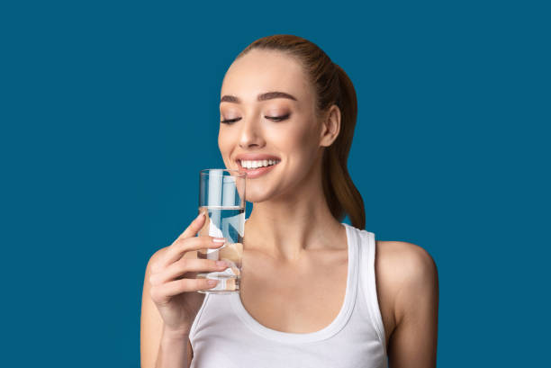 ragazza felice che beve vetro d'acqua in piedi su sfondo blu - bere acqua foto e immagini stock