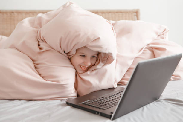 счастливая девушка, лежащая в постели под одеялом и учакласс виртуальной онлайн-школы. малыш работает на ноутбуке в интернете дома. ребенок - blue monday стоковые фото и изображения