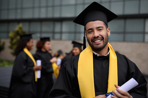 Portrait of a happy graduate student holding his diploma on graduation day and looking at the camera smiling - education concepts