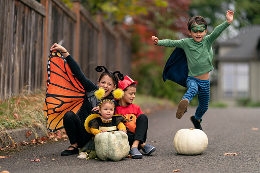 Four mixed race siblings in costumes happily play together outside in their driveway on Halloween. An elementary age boy dressed as a superhero is jumping over a large white pumpkin.