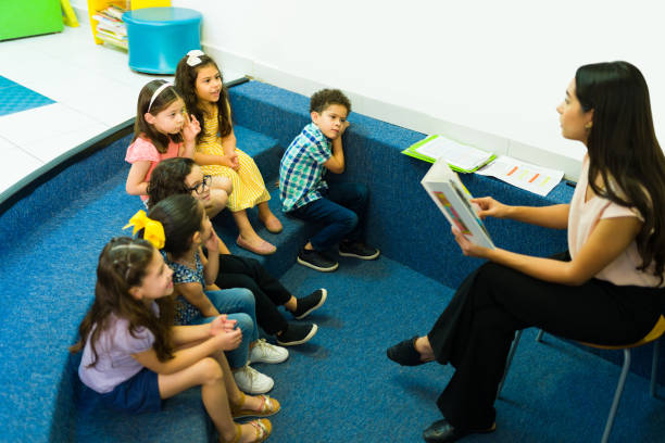 Happy group of preschoolers listening to a fairy tale Focused young kids enjoying listening to their kindergarten teacher reading a children's book teach reading to kindergarteners stock pictures, royalty-free photos & images