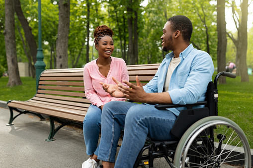 Happy handicapped black guy in wheelchair talking to his girlfriend at park, copy space. Young African American woman sitting on bench, communicating to her disabled male friend outdoors