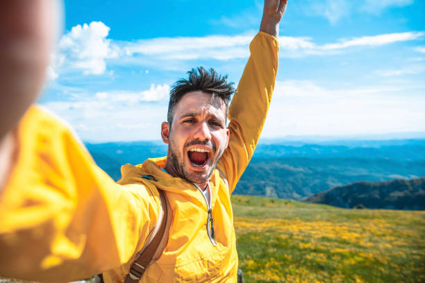 Happy hiker taking selfie portrait on the top of a mountain - Young man with arms up smiling at camera - Sport, people and technology concept stock photo