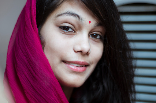 Beautiful Indian girl dressed in pink colored traditional Indian clothing and a headscarf stands in front of a dramatic sunset in pink and purple hues.  The portrait style headshot shows the girl wearing a big smile.  Copyspace. 