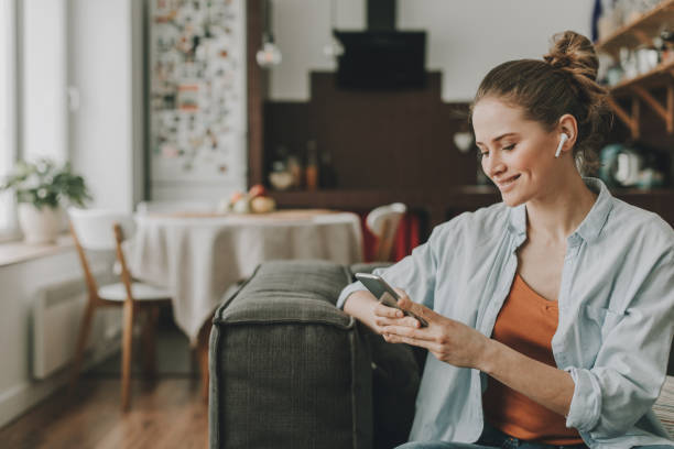 Happy lady sitting on couch with smartphone Technology in our life. Waist up portrait of young happy woman on sofa watching video on mobile phone with wireless earbuds bluetooth stock pictures, royalty-free photos & images