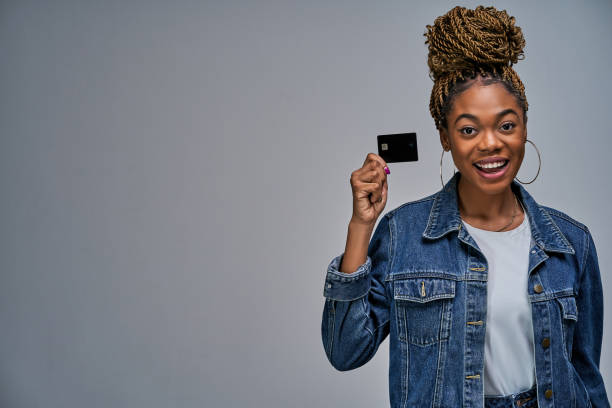 Happy lady with bun in a jeans jacket shows a black bank credit card in her hand. Banking concept A woman with a smile in earrings holding a credit card by fingers in hands. credit card photos stock pictures, royalty-free photos & images