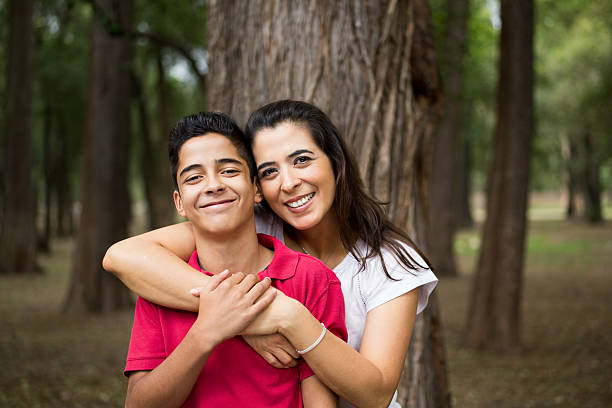 A happy latin mother and her teen son embracing and smiling at the camera in a horizontal waist up shot outdoors.