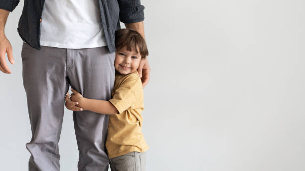 Happy little boy embracing his daddy, smiling to camera, feeling loved and safe, posing over grey wall with empty space stock photo