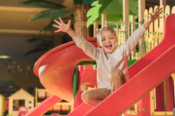 happy little boy smiling at camera while playing on slide in entertainment center happy little boy smiling at camera while playing on slide in entertainment center indoor playground stock pictures, royalty-free photos & images